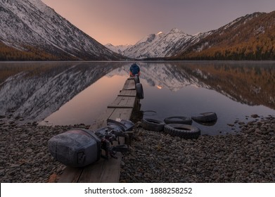 Dreamer. Lonely Man Greets The Dawn Sitting On A Boat Dock On The Shore Of Mountain Lake. Sunrise In Mountains. Old Boat Motor On A Wooden Pier.Reflection Of Snowy Peaks In Calm Water.Siberia, Russia - Powered by Shutterstock