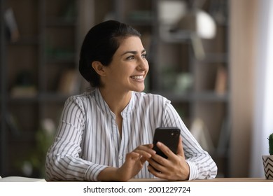 Dreamer with phone. Motivated indian female worker student sit at table look away hold cell think on good new career opportunities. Happy dreamy young lady inspired with job proposal received by email