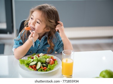 Dreamful Asian Girl Is Eating Fresh Tomato With Appetite. She Is Sitting And Looking Aside Playfully. Plate Of Salad And Glass Of Juice On Table. Copy Space