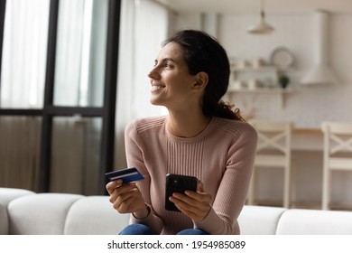 Dreamful latin woman shopper sit on couch at home look at distance with happy smile enjoy buying goods spending money. Young female plan future vacation book hotel tickets online use cell bank card
