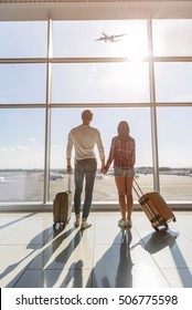 Dreamful Loving Couple Is Ready For Travel. They Are Standing Near Window At Airport And Looking At Flying Plane. Lovers Are Holding Hands