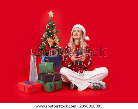 Dreamful Santa woman, full body view sitting near decorated Christmas tree dreamful Santa woman. Looking aside, hold use mobile cell phone. Pile of gift boxes, shopping bag. Celebrating alone.