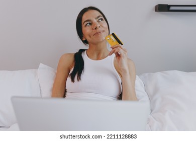 Dreamful Young Woman In White Tank Top Sit In Bed Using Laptop Pc Computer Holding Credit Bank Card Doing Online Shopping In Bedroom Lounge Home In Own Room House Wake Up Be Lost In Reverie Good Day