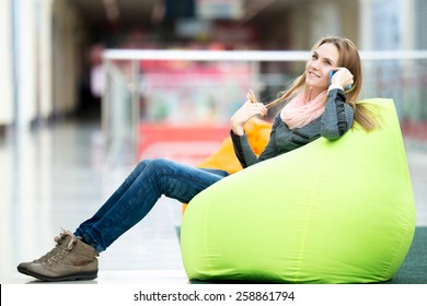 Dreamily Smiling Young Woman In Casual Style Clothes Sitting On Bean Bag In Contemporary Interior, Recreational Area, Using Smartphone, Talking, Making Call, Waiting For Meeting