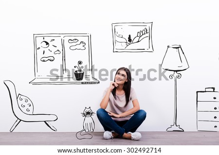 Dreaming about new house. Thoughtful young woman looking at the sketch on the wall while sitting on the floor 