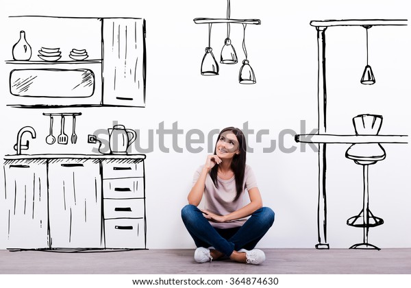 Dreaming about new kitchen. Cheerful young woman\
smiling while sitting on the floor against white background with\
drawn kitchen