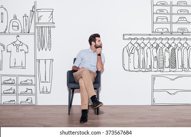 Dreaming About New Wardrobe. Young Handsome Man Keeping Hand On Chin And Looking Away While Sitting In The Chair Against Illustration Of Closet In The Background