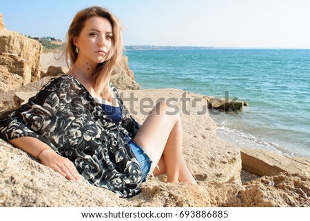 Dreaming beautiful girl with light hair dressed in shorts sitting lost in thoughts on big stones and looking forward. Rocks and sea on background.