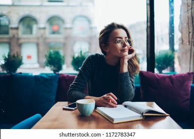 Dreaming beautiful hipster girl looking away while resting at cafeteria and writing romantic essay, attractive young woman in eyeglasses enjoying free time and planning to do list in notebook
