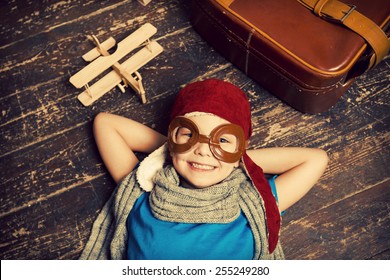 Dreaming of a big sky. Top view of happy little boy in pilot headwear and eyeglasses lying on the hardwood floor and smiling while wooden planer and briefcase laying near him 