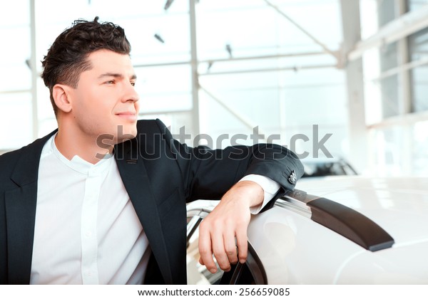 Dreaming of a new\
car. Smiling handsome man in formalwear leaning at the new car and\
looking away in car dealership\

