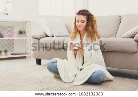 Dreaming redhead woman having rest. Wrapped up in white blanket and drinking coffee in warm atmosphere, sitting on floor at home, copy space