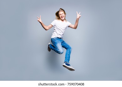Dreams come true! Cheerful glad and funky guy with long blonde hair dressed in casual clothes is jumping up and demonstrating v-sign, isolated on grey background