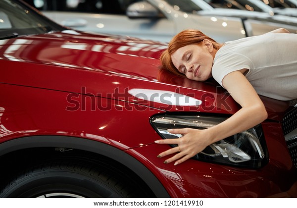 Dreams come true today. Young emotive\
cute woman with amazed expression, embracing her new cargently at\
car show room. Husband bought her new car for\
b-day.