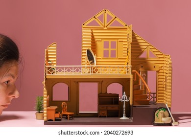 Dreams Of Owning Your Own House, Choosing Real Estate, A Fun Concept. A Young Woman Looks Like A Giant At A Small Two-story House. Dollhouse On A Pink Background, Toy Furniture Inside.