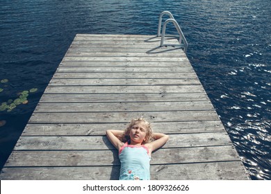 Dreams And Relaxation. Girl Child With Closed Eyes Lying On Wooden Lake Dock On Sunny Day. Kid Dreaming Enjoying Summer Outdoor. Happy Childhood Lifestyle. View From Above Overhead.