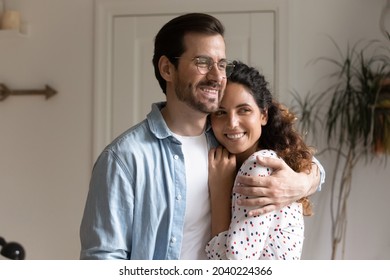 Dreamy 30s Couple Standing Indoor Hugging Looking Out Window Daydreaming. Husband And Wife First Time Home Buyers Homeowners Feels Happy, Indulge In Dreams About Bright Future Life Together Concept