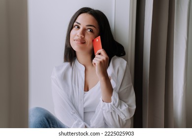 Dreamy African American Woman Sitting On Windowsill Looking At Camera Holding Credit Card, Wearing White Shirt And Blue Jeans At Home. Satisfied Brazilian Girl Earned Money To Buy A New Home