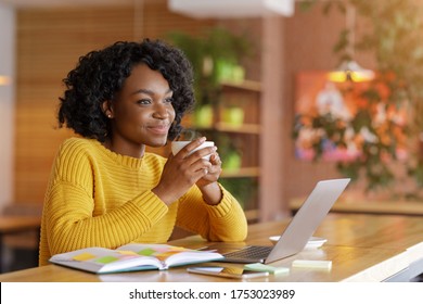 Dreamy african girl dinking tea at cafe while looking for job online, using laptop and taking notes, copy space