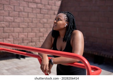 Dreamy Afro American Woman In Black Sportive Clothes Having Rest, Looking At Side In City, In Sports Ground, Relaxing, Thinking, Having Some Minutes To Take A Break During Jogging Or Fitness Workout