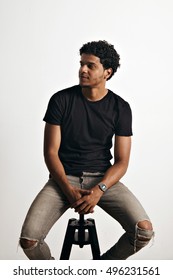Dreamy athletic relaxed black young man wearing torn jeans and an unlabeled black t-shirt sitting on a small stool against white wall background