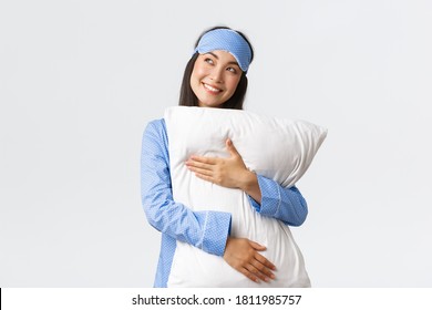 Dreamy beautiful asian girl in blue pajamas and sleeping mask hugging pillow and smiling pleased as looking upper left corner imaging something in bed, picturing, using imagination - Powered by Shutterstock