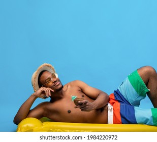 Dreamy Black Man Sunbathing On Inflatable Lilo, Drinking Tropical Cocktail From Coconut Shell On Blue Studio Background, Copy Space. Cheerful Young Guy Chilling On Beach, Enjoying Fresh Beverage