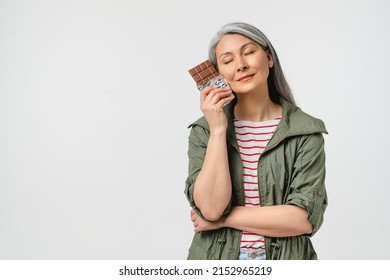 Dreamy Caucasian Mature Middle-aged Woman With Grey Hair In Casual Clothes Wants To Eat Chocolate Bar Full Of Calories Isolated In White. Eating Disorder Concept. Junk Fat Unhealthy Food. Dieting