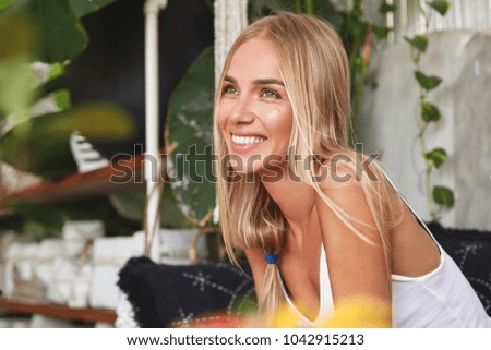 Dreamy cheerful positive young female with blonde hair, looks somewhere into distance, sits on comfortable sofa, thinks about something pleasant or dreams to have unforgettable summer recreation
