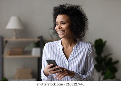 Dreamy cheerful young african american multiracial woman holding smartphone in hands, feeling excited of getting message email with pleasant news, visualizing future or daydreaming alone at home.