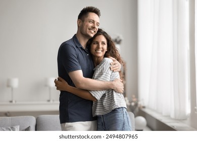 Dreamy couple in love standing in modern living room looking out window and smiling, enjoy date, dreaming about bright future together at their new bought house. Relations, happy marriage, daydreams - Powered by Shutterstock