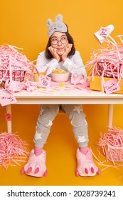 Dreamy Cute Asian Student Dressed In Soft Pajama And Sleepmask On Forehead Has Breakfast At Workplace Enjoys Cozy Home Environment Poses At Messy Desktop With Paper Rubbish Yellow Background