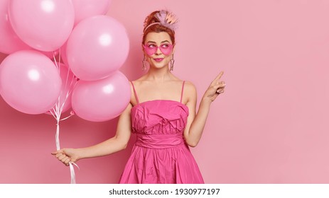 Dreamy fashionable woman wears sunglasses and festive dress points away on blank space holds bunch of inflated balloons enjoys holiday isolated over pink background. Special occasion concept - Powered by Shutterstock