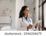 Dreamy female doctor, professional medical worker using digital tablet in clinic, looks out window with smile, web surfing information, giving distant healthcare advice, modern technology and medicine