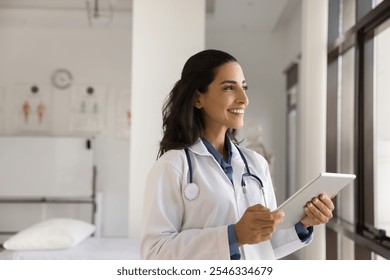 Dreamy female doctor, professional medical worker using digital tablet in clinic, looks out window with smile, web surfing information, giving distant healthcare advice, modern technology and medicine - Powered by Shutterstock