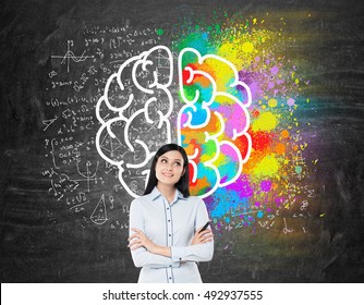 Dreamy girl with black hair standing near blackboard with brain sketches on it. Concept of brain studying