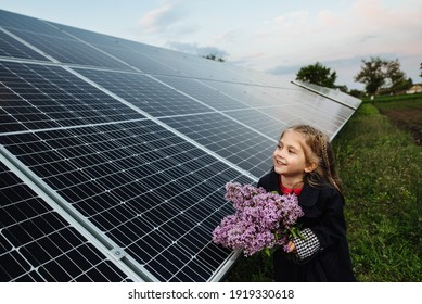 Dreamy girl with a bouquet of flowers on a background of solar panels. Portrait of a smiling child holding lilac flowers in the area near the house. The concept of ecology, a happy future. Happy child - Shutterstock ID 1919330618