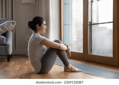 Dreamy Indian woman looking out window, sitting on warm wooden floor at home, pensive young female in grey sportswear dreaming of good future in living room, visualizing success, new opportunities - Powered by Shutterstock