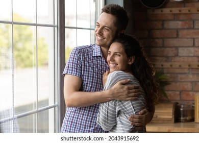 Dreamy Loving Young Husband Cuddling Happy Beautiful Hispanic Wife, Visualizing Future Looking In Distance Out Of Window, Enjoying Tender Sweet Family Moment Or Celebrating Moving Into Own House.