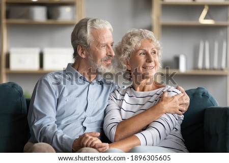 Dreamy middle aged senior loving retired family couple looking in distance, planning common future or recollecting memories, enjoying peaceful moment relaxing together on cozy sofa in living room.