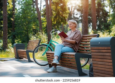 Dreamy modern old lady relaxing city park. Pensive senior gray haired woman casual sitting wooden bench outdoors reading book. cycling forest park, bicycle, healthy active lifestyle after 50-60 years