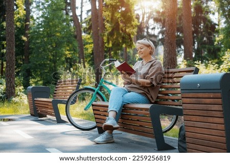Dreamy modern old lady relaxing city park. Pensive senior gray haired woman casual sitting wooden bench outdoors reading book. cycling forest park, bicycle, healthy active lifestyle after 50-60 years