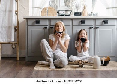Dreamy mother sitting on kitchen floor together with her lovely daughter, enjoying morning coffee, drinking beverage and spending weekend in cozy apartment, resting at home
