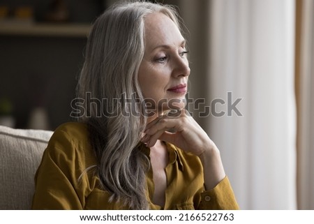 Dreamy peaceful aged female rest sit on sofa looks aside feel carefree spend leisure alone at home, deep in pleasant memories and thoughts. Contemplation, relax, older beautiful woman portrait concept