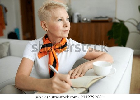 Dreamy pensive elderly female novelist sitting on couch in living-room leaning on its back and writing new story, inventing breathtaking plot and heroes of novel, looking aside with thoughtful face