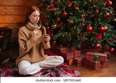 Dreamy pensive redhead young woman wearing winter sweater writing Christmas letter to Santa Claus sitting on floor at home on background of xmas tree at cozy living room with festive interior.