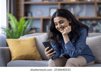 A dreamy and pensive young Indian girl is sitting on the sofa at home, resting her head on her hand and smilingly looking at the mobile phone screen. - Powered by Shutterstock