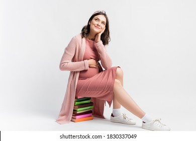 dreamy and pregnant woman sitting on books and looking away on white