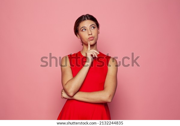 Dreamy pretty cute woman touching\
her chin thought choose decide solve problems dilemmas wearing\
fashionable outfit on pink background. Youth people portrait.\
