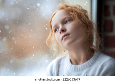 A dreamy red-haired little girl in white sweater looking pensively out the window. Natural lighting. Children, childhood, emotions. Winter season. Copy space. - Powered by Shutterstock